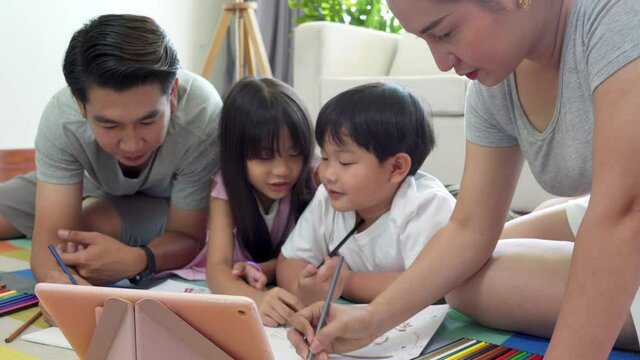 Happy Asian family at home. Parents with little daughter and son lying on the floor with using color pencil painting on coloring book. Father and mother with two kids having fun homeschooling together