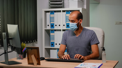 Entrepreneur with mask typing on computer sitting in new normal workplace. Freelancer working in modern office workspace cleaning hands using sanitize alcohol gel against corona virus