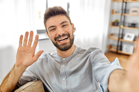 technology, people and lifestyle concept - happy man taking selfie or having video call and waving hand at home