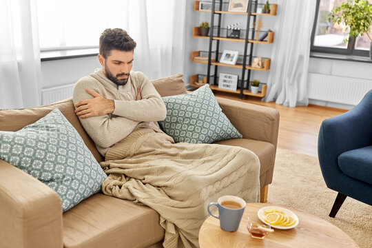 health, cold and people concept - sick young man in blanket drinking hot tea with lemon and honey at home