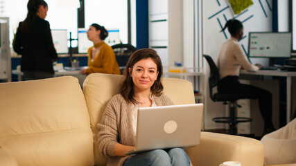 Young woman entrepreneur sitting on couch in middle of business start up office looking at camera smiling holding laptop. Multiethnic coworkers talking about startup financial company in modern office © DC Studio