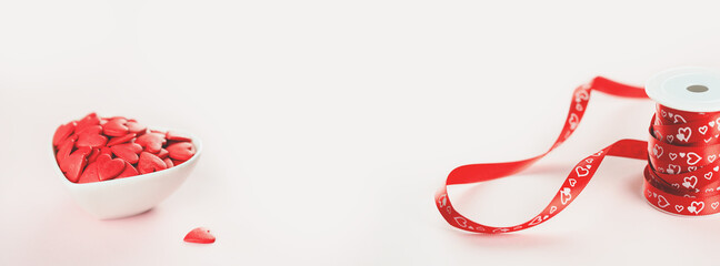 Heart shaped red sprinkles in a white bowl and gift ribbon roll, festive love gift and Valentine's Day concept or wedding day. wide banner. Small hearts. Selective focus