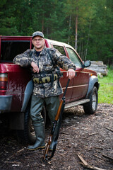 Man hunter standing with shotgun rifle near his car outdoor preparing for bird hunt in autumn forest