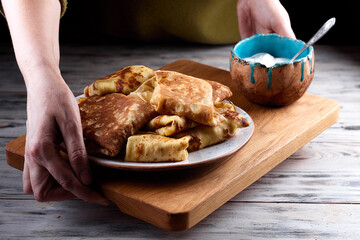 Homemade crepes, tasty thin pancakes on plate with a bowl of sour cream on old wooden plank in female hands