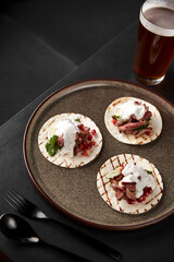 Fototapeta na wymiar Vertical frame in dark colors. Delicious appetizer of meat, onions, pomegranate seeds on a wheat tortilla with white sauce and a glass of light beer on a black table