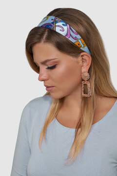 Blonde European lady in pale blue jumper and with golden chandelier earrings is looking down. The girl's hair is fixed with blue silk headband with multicolor abstract print. 