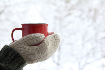 a red mug in the hands of those dressed in knitted mittens against the backdrop of a blurred snow...