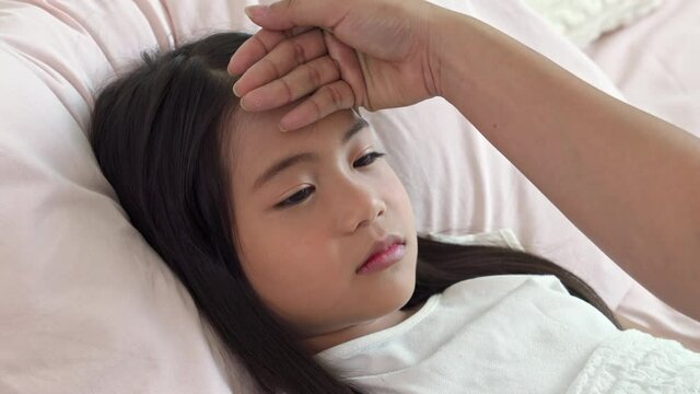 Asian woman using hand checking temperature on sick daughter forehead. Mother put fever patch on illness cute child girl forehead on the bed in bedroom. Family and medical health insurance concept.