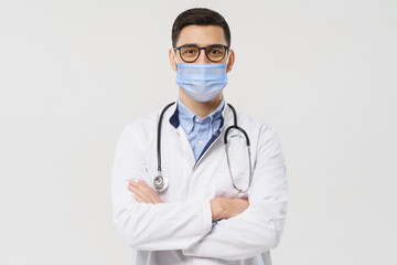 Portrait of youn doctor wearing surgical mask, holding crossed arms, ready to help patients with coronavirus or covid virus, isolated on gray background