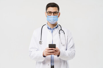 Portait of young doctor wearing surgical mask and white coat, holding his phone with both hands, using medical app, isolated on gray background