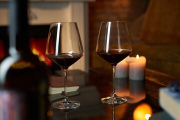 Two glasses of red wine and candle on table at home, fireplace in the background. Warm, dark colors.
