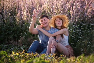 Naklejka na ściany i meble Happy couple in love sits hugging by blooming sage field. A young man with beautiful girl in straw hat are having fun, fooling around, waving their hands, showing their tongue. travel of crazy lovers
