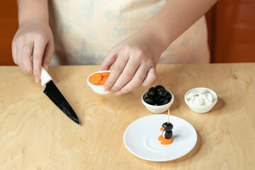 Woman’s hands preparing a healthy food
ingredients for making a skewer snack made from olives, curd cheese and carrots in the shape of a penguin