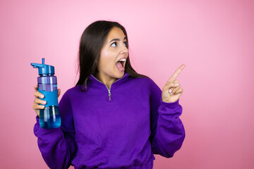 Young beautiful woman wearing sweatshirt holding a water bottle over isolated pink background smiling and pointing with hand and finger to the side