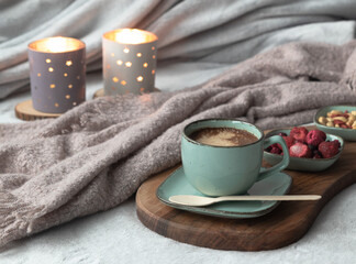 Fototapeta na wymiar Cappuccino coffee in a green mug, biodegradable eco spoon, berries and nuts on dark wooden cutting board on grey soft cozy hygge plaid blanket. Burning candles