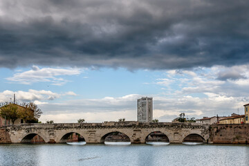 The ancient Bridge of Augustus and Tiberius is reflected in the water of the canal in the historic center of Rimini, Italy, under a dramatic sky