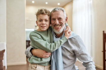 Happy grandfather embracing with his cute little preschool grandson, standing together at home and smiling at camera - obrazy, fototapety, plakaty