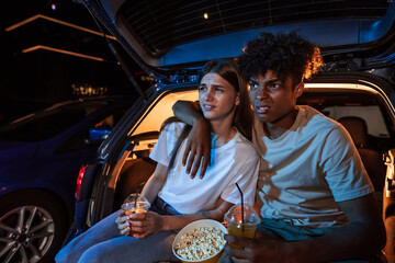 Beautiful young couple embracing, watching a movie, having popcorn while sitting together in car trunk in front of a big screen in an open air cinema