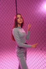 Fashion model in green sporty costume. Pretty girl with long straight hair standing and posing against grid fence and pink background