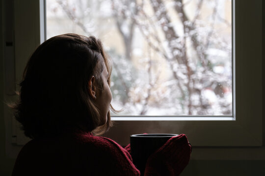 Young Woman Holding A Hot Coffee Or Tea Cup Looking Out Of The Window In A Snowy Winter Day. Cozy At Home Concept.