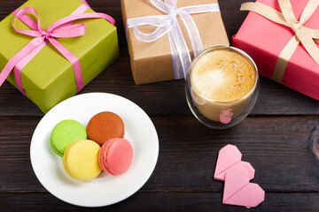 White plate with French macarons, two gift boxes, small paper hearts and a glass of latte. A gift for a loved one.