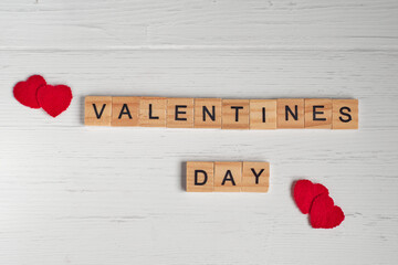Top view of the word valentines day laid out from square wooden tiles and red small hearts on old white wooden background.