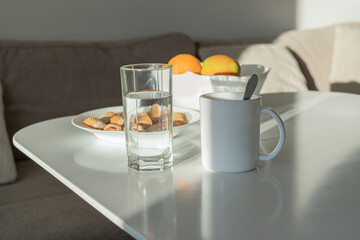 Water or coffee? Which is better? A white cup and a glass of water stand on a bright table. In the background are fruits and cookies.