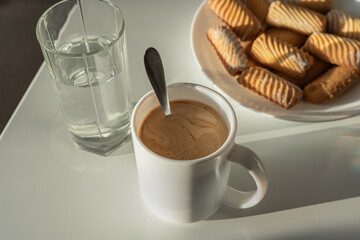 A white cup with coffee and a teaspoon stands on a bright table. In the background is a glass of water and cookies.