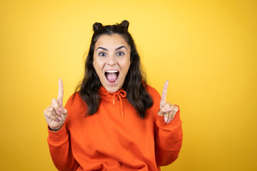 Young beautiful woman wearing sweatshirt over isolated yellow background amazed and surprised looking at the camera and pointing up with fingers and raised arms