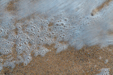 Close up sea water waves with bubbles on sand beach. Pattern and texture surface with white and orange colors. Abstract outdoor natural view as a background.