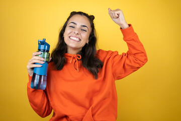 Young beautiful woman wearing sweatshirt holding a water bottle over isolated yellow background showing arms muscles smiling proud