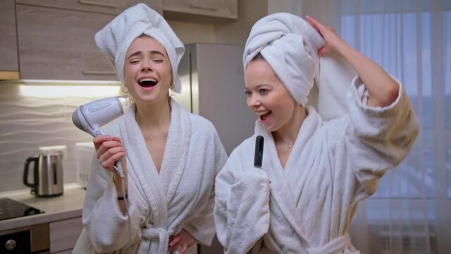 Two girl friends in white bathrobe dance, sing and dry hair with hairdryer, laugh. Young women have fun time. Funny video