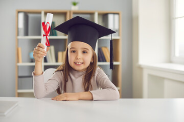 Education for children concept. Adorable preschool girl in academic cap looking at camera, holding kindergarten graduate diploma. Webcam view kid in mortarboard sitting at desk in classroom or at home