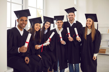 Group portrait of smiling multicultural university or college graduates standing together, holding diplomas and looking at camera. Happy students in traditional caps and robes celebrating graduation