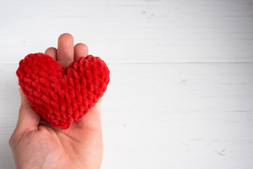 Male Caucasian hand holding a red knitted heart on an old white wooden background with copy space for text. The concept of love, valentine's day, care.