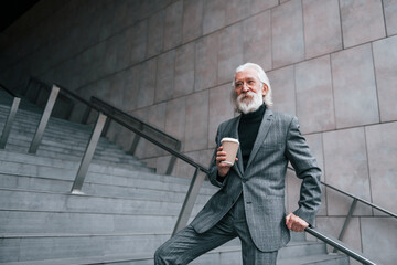 Senior businessman in formal clothes, with grey hair and beard is outdoors and holds cup of drink