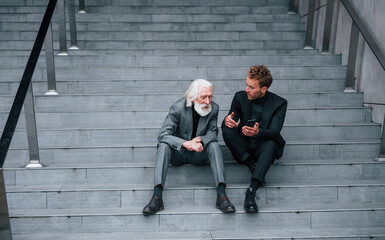 Sitting with phone. Young guy with senior man in elegant clothes is outdoors together. Conception of business