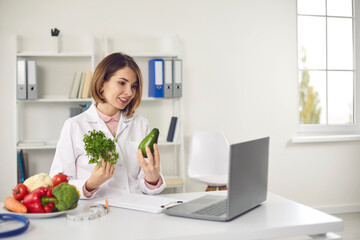 Obraz na płótnie Canvas Young woman doctor nutritiologist showing fresh ingredients to patient online on laptop and speaking about healthy balanced diet during videlcall or distant meeting. Healthy lifestyle concept