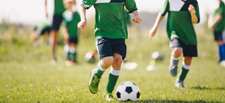 Kids Running After Soccer Ball At Natural Grass Sports Field. Group Of Children Training Football Skills At Physical Education Class. Sports Summer Practice For Kids With Coach