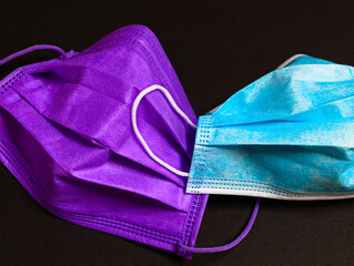 Eine violette und eine blau-weiße OP-Maske vor schwarzem Hintergrund 