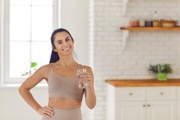 Naklejka na ściany i meble Drink water. Lifestyle portrait of a beautiful slender fitness woman holding a glass of water while standing in the kitchen. Concept of good habits, hydration, diet and sports. Banner.