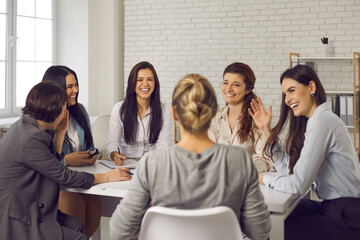 Team of happy young business women sitting around office table, discussing ideas, sharing funny stories and laughing. Group of smiling company employees talking and having fun in corporate meeting