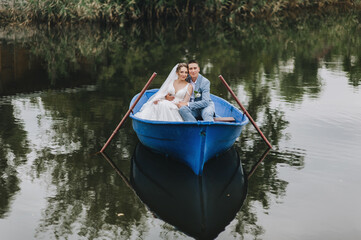 A loving groom in a suit and a cute bride in a white lace dress are sitting in a wooden boat, walking and swimming on the lake, reflected in the water, enjoying the beautiful nature. Wedding portrait.