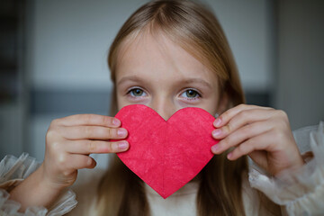 Little school kid girl holding red heart. Valentine's day during pandemic coronavirus covid-19 quarantine. Child showing heart online via video chat to boy friend