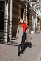 Portrait of a young woman in coat with closed eyes standing on street in front of window and brown wall, enjoying sunlight. Caucasian girl having fun outside. Urban style and fashion. Casual outfit