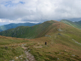 Lonely hiker walking e at hiking trail at grassy mountain ridge, hill slopes at bad weather with strong wind, thick fog and clouds, Chopok, Low Tatras, Slovakia, late summer day