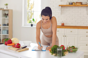 Cheerful pretty slim female athete in sportswear standing in kitchen in sportswear and writing down healthy recipe or daily ration diet at home. Active healthy lifestyle, clean eating concept - obrazy, fototapety, plakaty