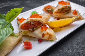Banquet serving of pancakes with red caviar and salmon, on a white rectangular plate, with basil leaves and an orange slice