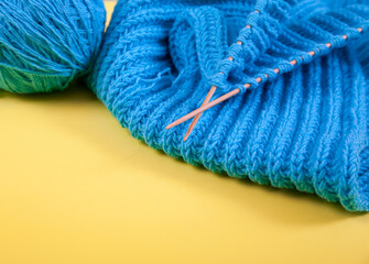A rolled up hand-knitted blue scarf, balls of yarn and knitting needles lie on a yellow background.