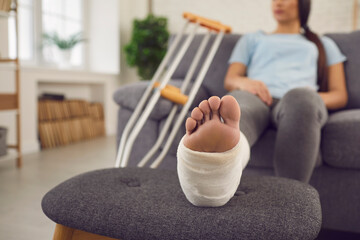 Woman with broken leg sitting on sofa with crutches. Young patient with serious bone fracture...